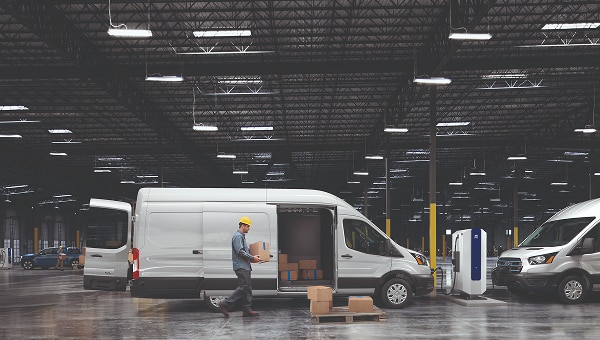 Trabajador cargando una Ford E-Transit blanca en un almacén, con otra van eléctrica cargándose al lado.